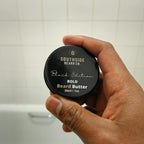 Hand holding a Southside Beard Co. bold beard butter container against a tiled bathroom wall.