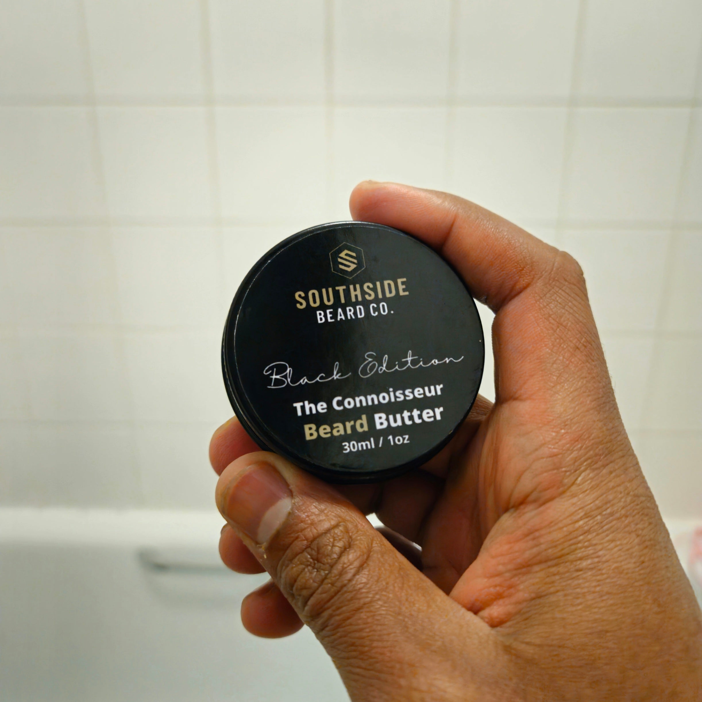 Hand holding a container of Southside Beard Co. The connoisseur beard butter against a tiled bathroom wall.